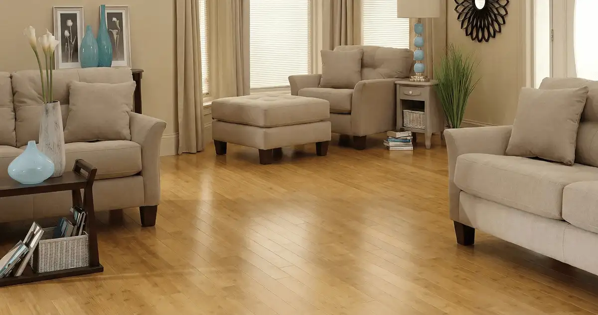 A living room with professionally installed bamboo flooring, beige sofas, a matching ottoman, a side table with a blue lamp, vases, framed pictures, and large windows with blinds and curtains in Baltimore, MD